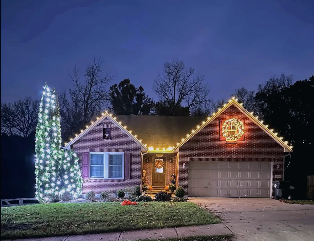 Warm white roofline with a lit wreath and a fully wrapped evergreen at dusk