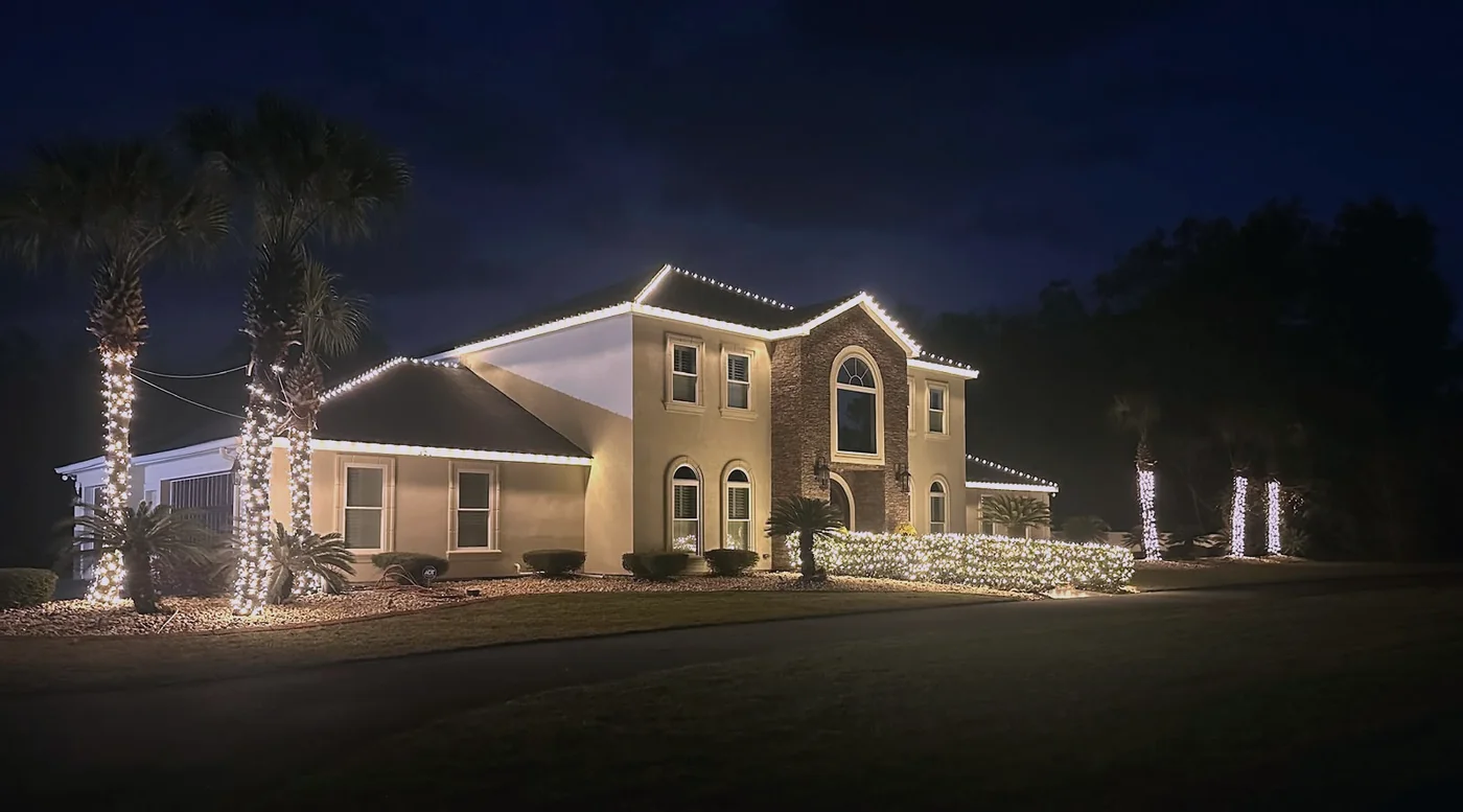 Florida home at dusk with warm white roofline, wrapped palms and lit hedge